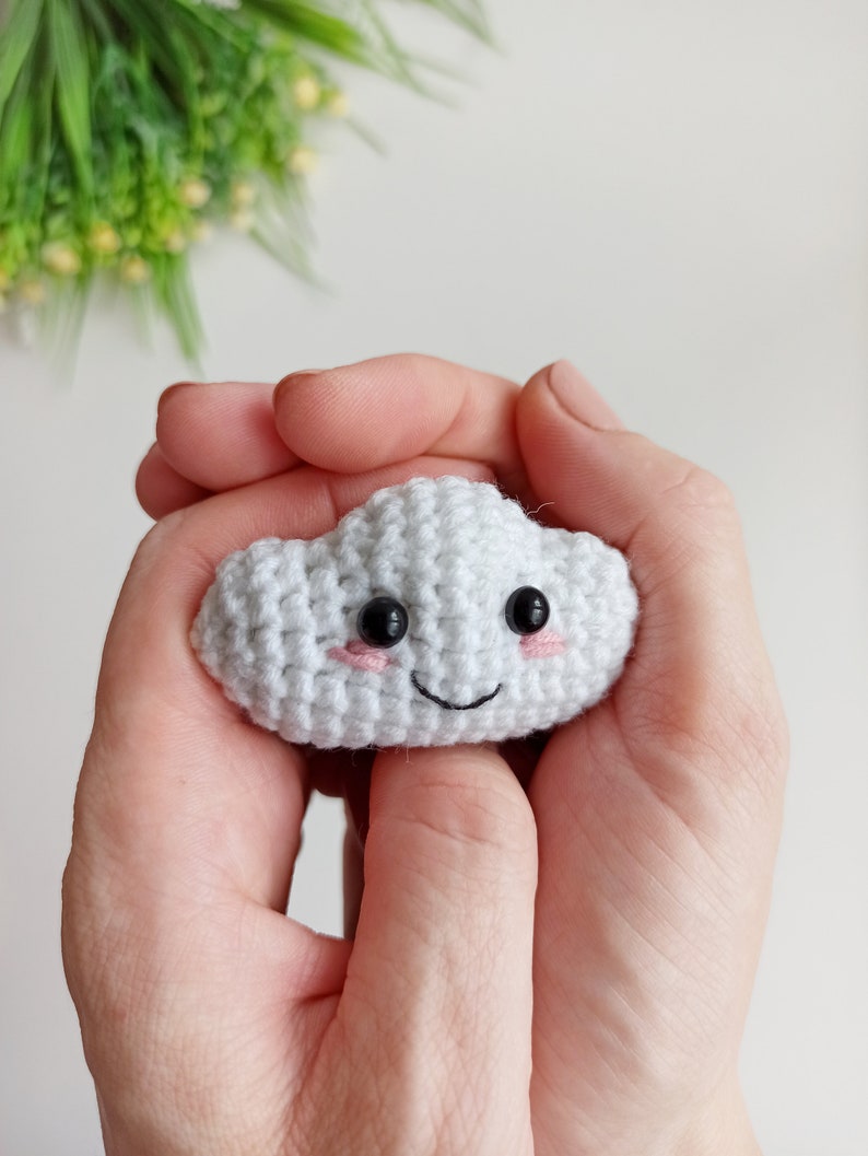 May include: A white crocheted cloud with a smiling face, black eyes, and pink cheeks. The cloud is being held in a person's hands.