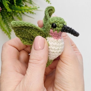 May include: A small crocheted hummingbird with green wings, a white chest, and a pink throat. The hummingbird has black eyes and a black beak.