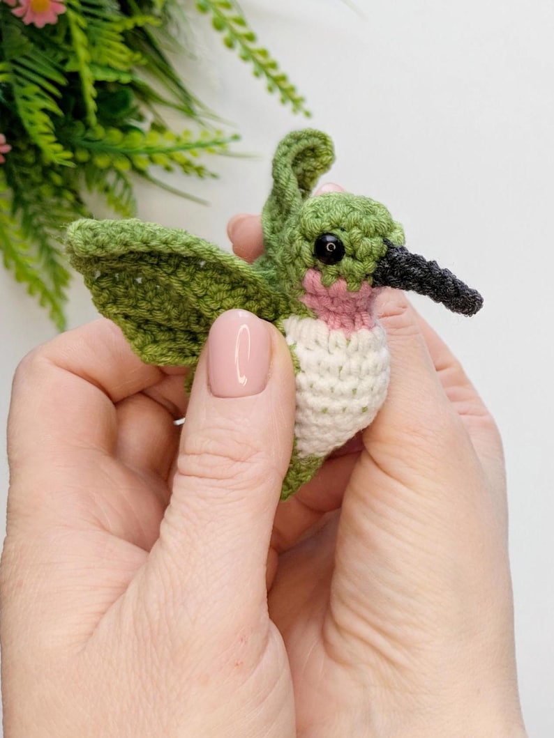 May include: A small crocheted hummingbird with green wings, a white chest, and a pink throat. The hummingbird has black eyes and a black beak.