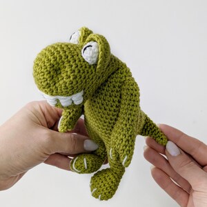 Puede incluir: Un juguete de dinosaurio verde tejido a mano con detalles blancos. El dinosaurio tiene una boca sonriente con dientes blancos, ojos blancos y costuras blancas en los pies. El juguete est&aacute; sostenido por dos manos sobre un fondo blanco.
