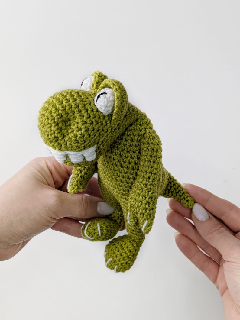 Puede incluir: Un juguete de dinosaurio verde tejido a mano con detalles blancos. El dinosaurio tiene una boca sonriente con dientes blancos, ojos blancos y costuras blancas en los pies. El juguete est&aacute; sostenido por dos manos sobre un fondo blanco.