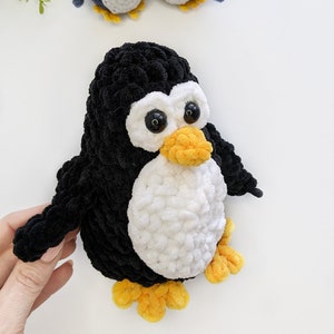 Pode incluir: Um pinguim de croch&ecirc; preto e branco com p&eacute;s laranja e grandes olhos pretos. Dois pinguins de croch&ecirc; azuis menores est&atilde;o no fundo.
