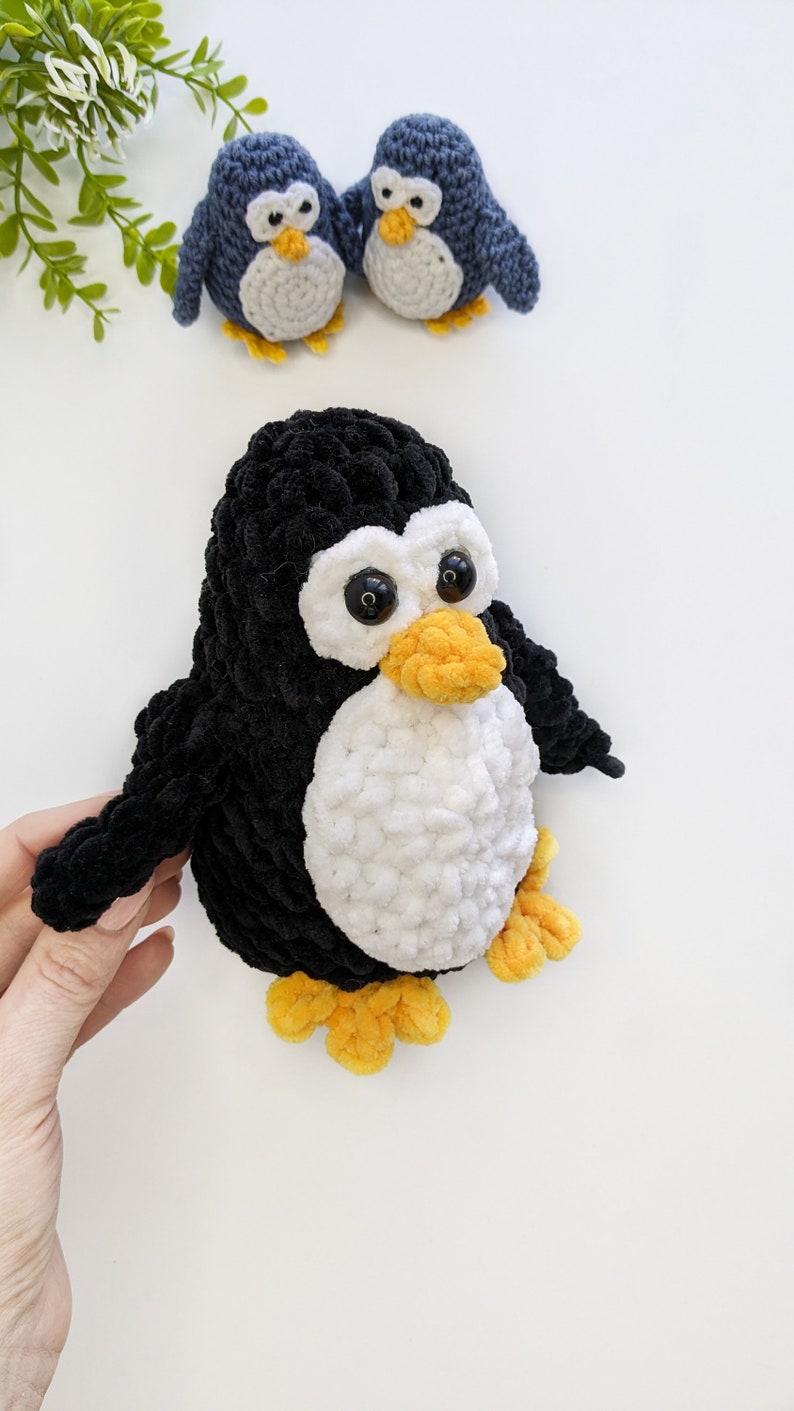Pode incluir: Um pinguim de croch&ecirc; preto e branco com p&eacute;s laranja e grandes olhos pretos. Dois pinguins de croch&ecirc; azuis menores est&atilde;o no fundo.