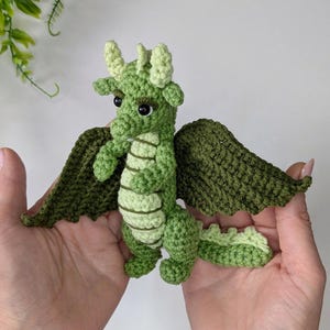 May include: A green crocheted dragon with wings, a long tail, and large eyes. The dragon is being held in two hands.