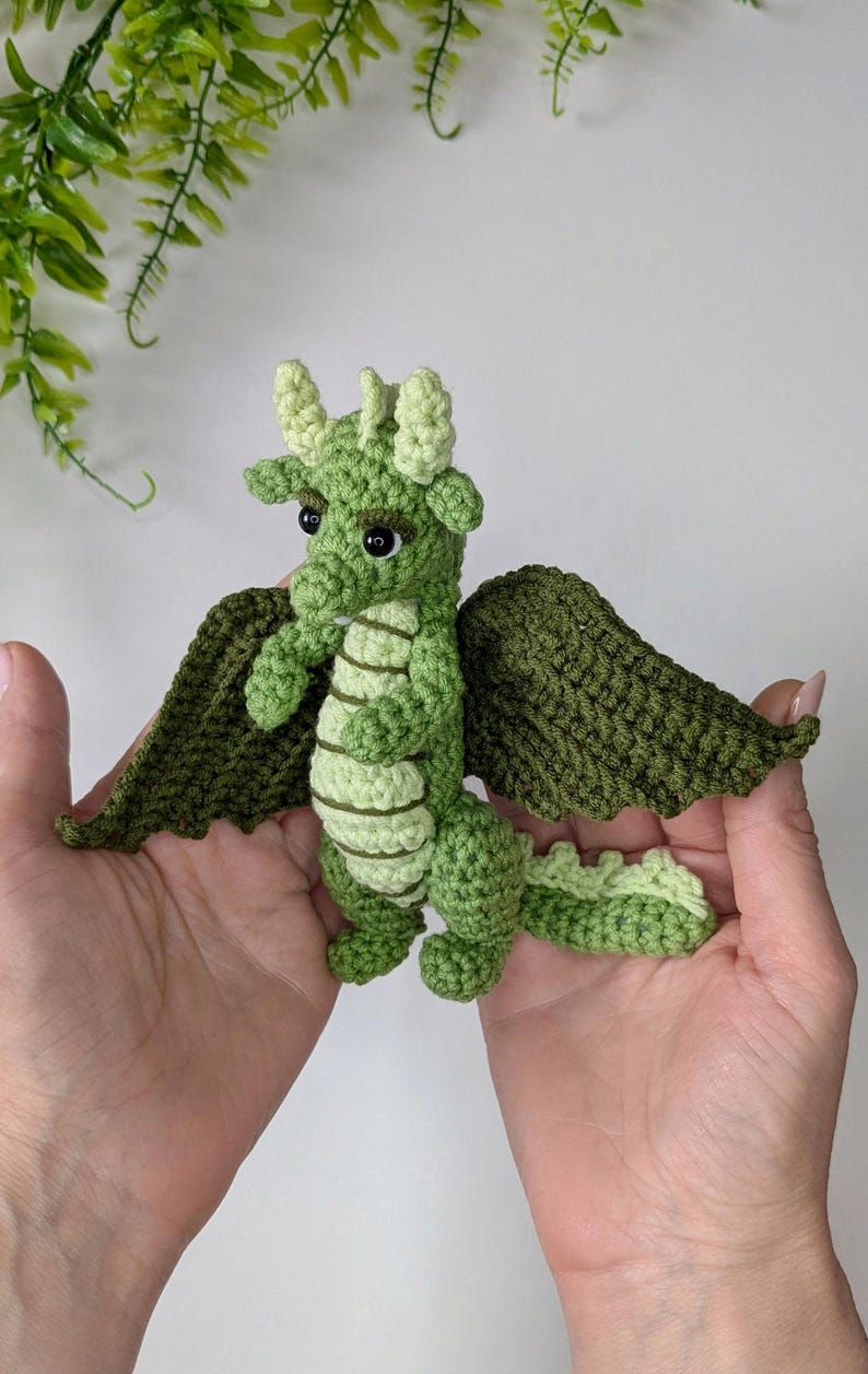 May include: A green crocheted dragon with wings, a long tail, and large eyes. The dragon is being held in two hands.