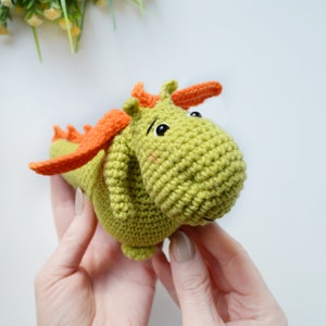 May include: A green crocheted dragon toy with orange wings and black eyes. The dragon is being held by two hands.