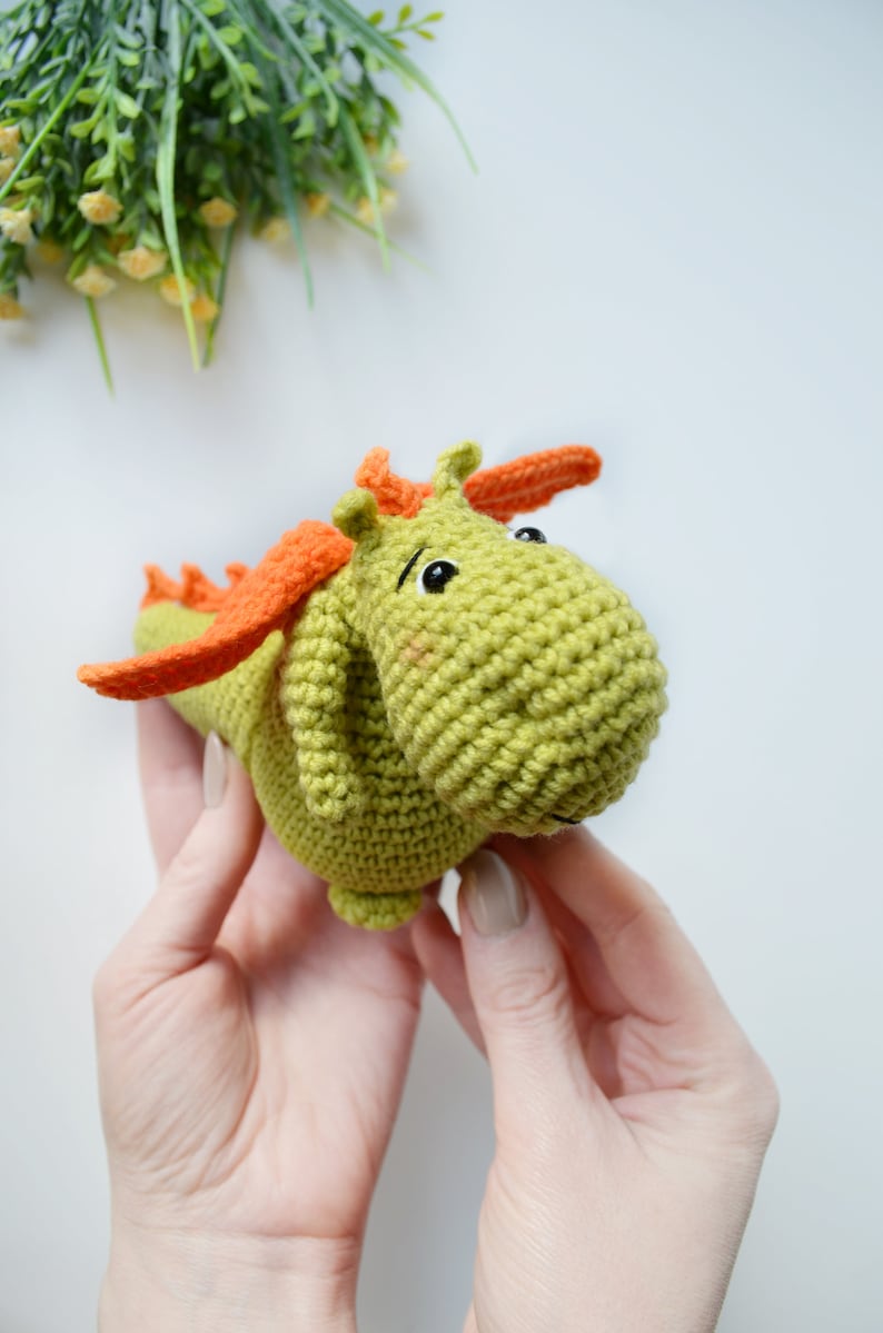 May include: A green crocheted dragon toy with orange wings and black eyes. The dragon is being held by two hands.