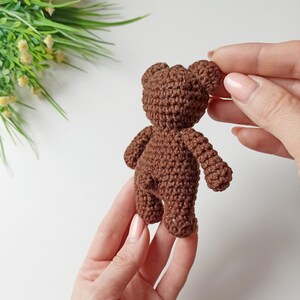 May include: A small brown crocheted teddy bear with a simple design. The bear is being held by a person's hands.