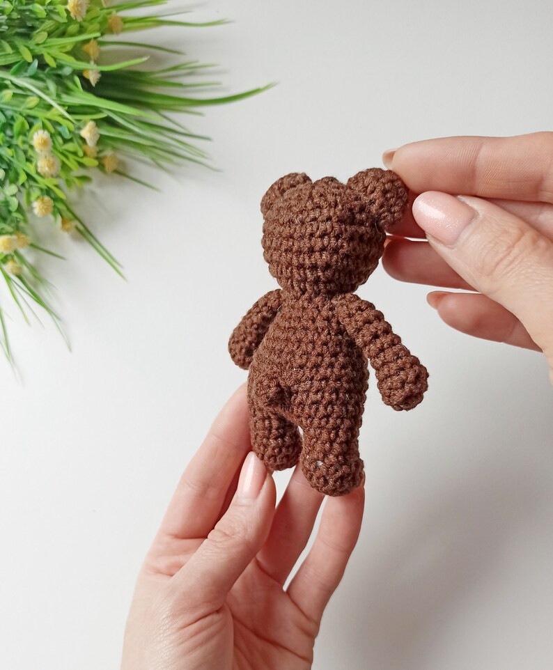 May include: A small brown crocheted teddy bear with a simple design. The bear is being held by a person's hands.