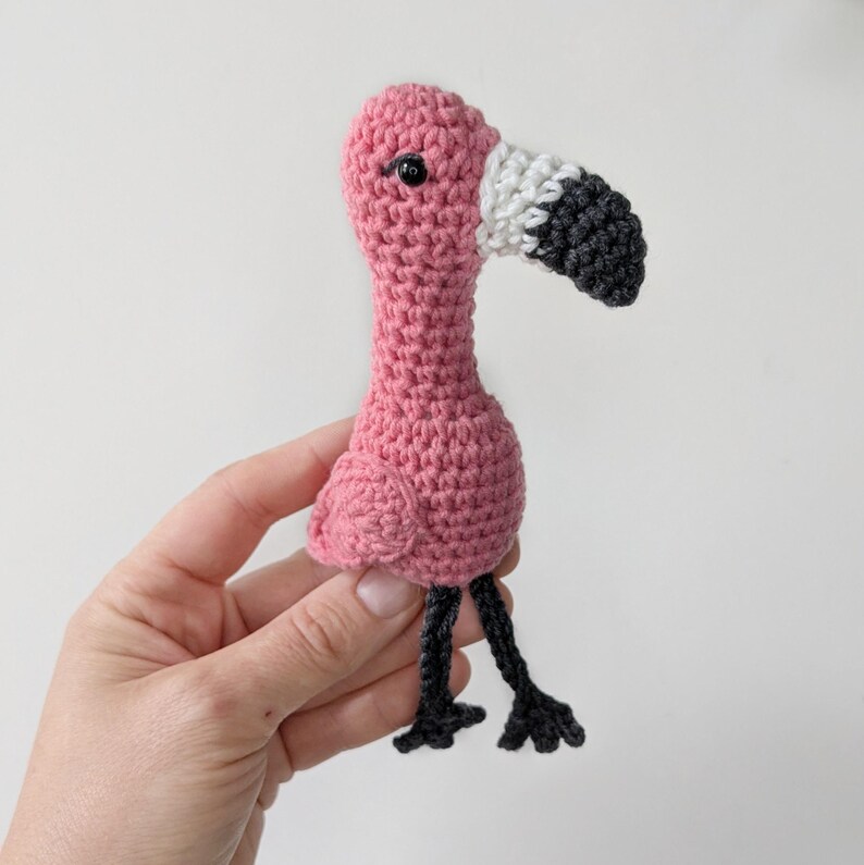 May include: A small, handmade crochet flamingo toy featuring a bright pink body, a white and dark gray curved beak, and two thin, dark gray dangling legs. The flamingo has a single black bead eye and a textured, yarn-based construction.