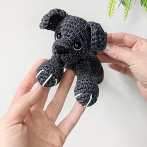 K&ouml;nnte beinhalten: Ein kleines, dunkelgraues H&auml;kelspielzeug in Form eines Hundes mit Schlappohren, schwarzen Kunststoffaugen und einer schwarz gestickten Nase. Die Vorderpfoten haben wei&szlig;e Stickdetails als Krallen, gefertigt aus strukturiertem Garn.