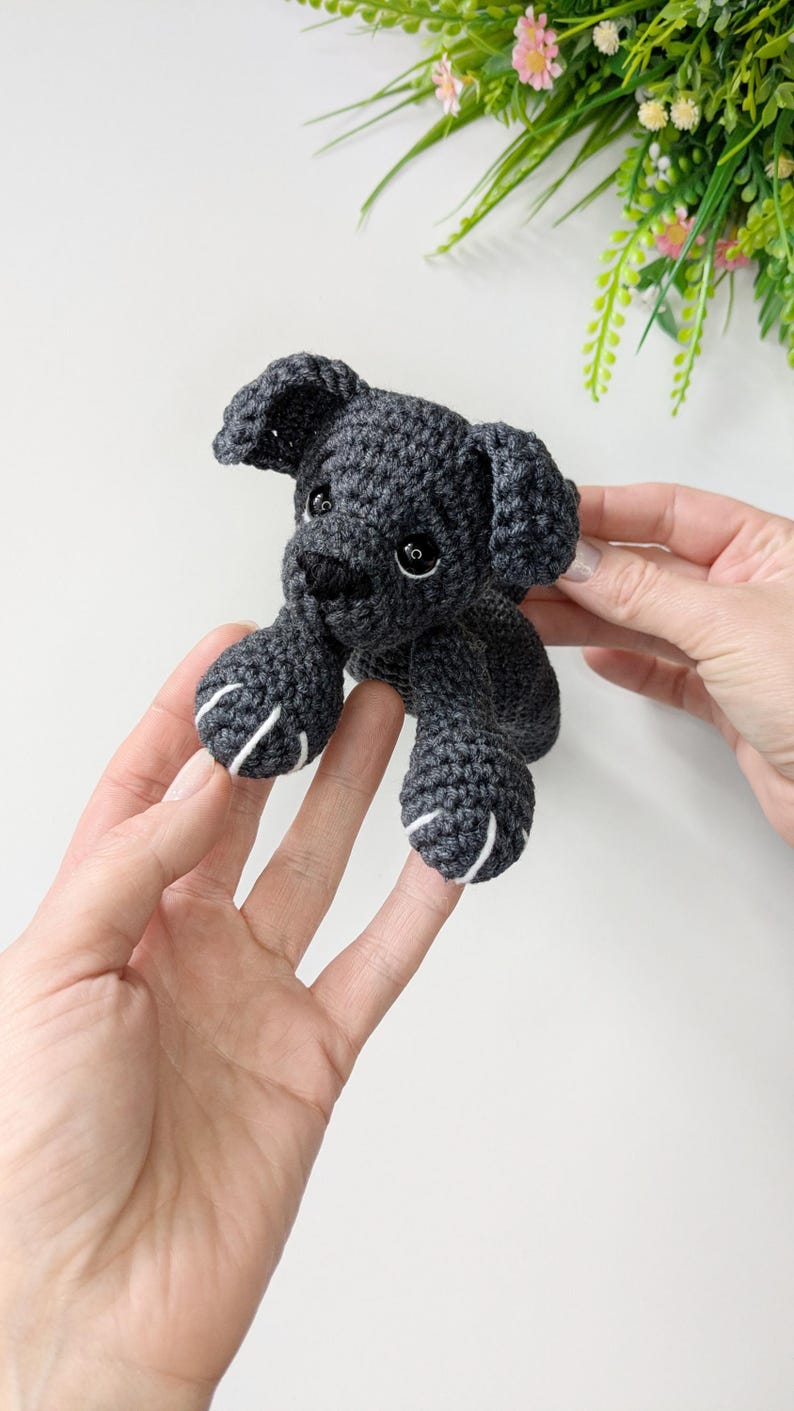 K&ouml;nnte beinhalten: Ein kleines, dunkelgraues H&auml;kelspielzeug in Form eines Hundes mit Schlappohren, schwarzen Kunststoffaugen und einer schwarz gestickten Nase. Die Vorderpfoten haben wei&szlig;e Stickdetails als Krallen, gefertigt aus strukturiertem Garn.