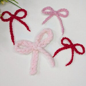 May include: Four handmade yarn bows in shades of red and pink. Two bows are a vibrant red, while the other two are soft pink. The bows are arranged on a white surface, showcasing their textured, fluffy appearance. These bows are perfect for crafting.