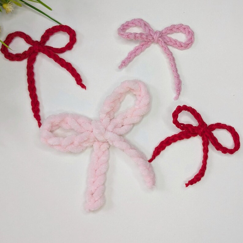 May include: Four handmade yarn bows in shades of red and pink. Two bows are a vibrant red, while the other two are soft pink. The bows are arranged on a white surface, showcasing their textured, fluffy appearance. These bows are perfect for crafting.