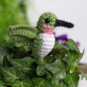 May include: A crocheted hummingbird made with green, white and pink yarn, perched on a green leafy plant in a blue and white patterned pot.