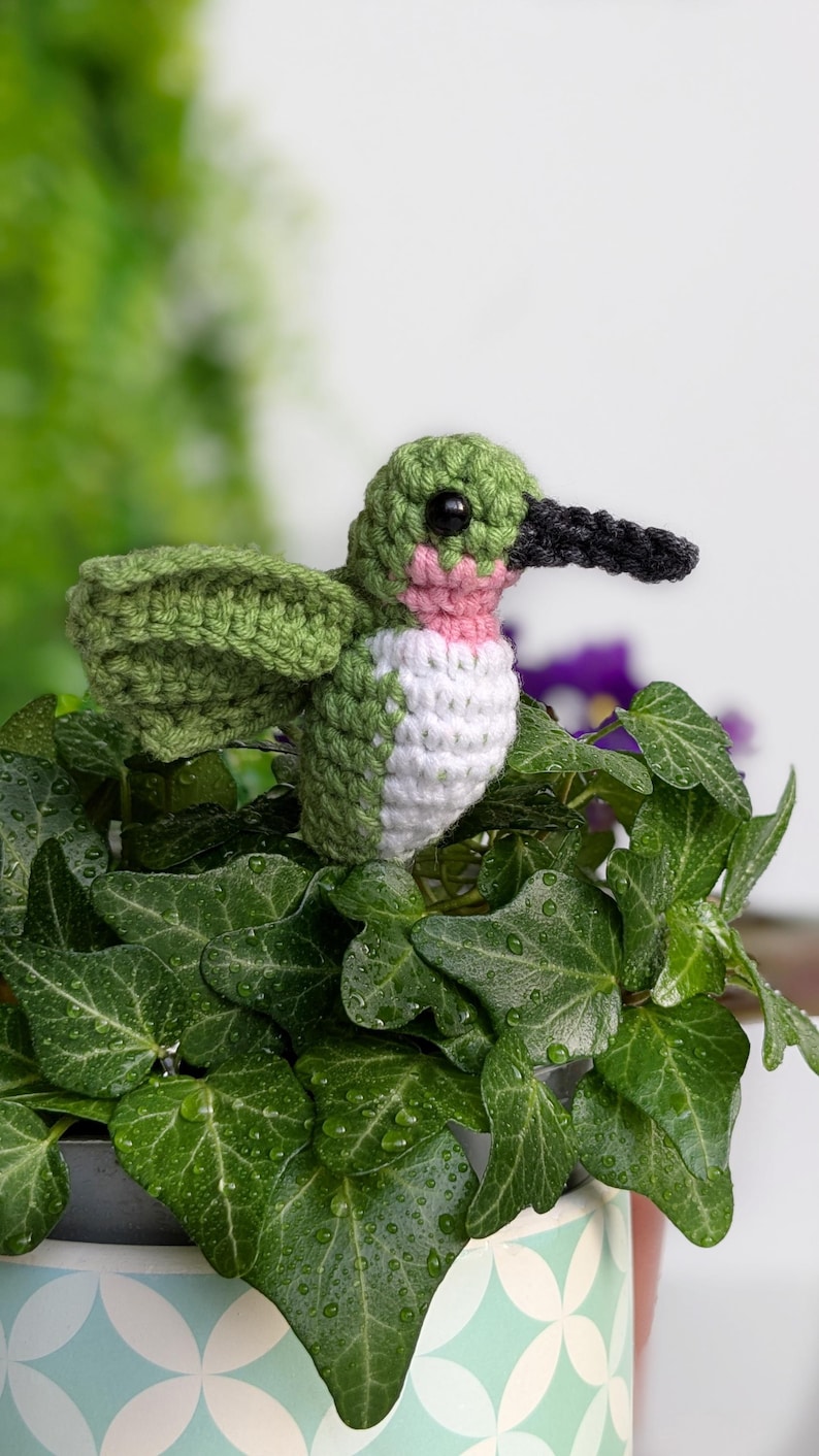 May include: A crocheted hummingbird made with green, white and pink yarn, perched on a green leafy plant in a blue and white patterned pot.