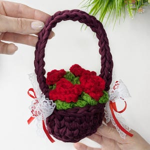 May include: A handmade burgundy crochet basket with a braided handle, filled with red crochet roses and green leaves. White lace and red ribbon accents adorn the sides. A charming, decorative floral basket.
