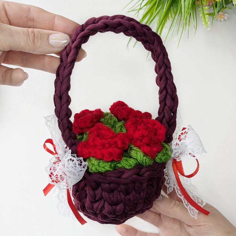 May include: A handmade burgundy crochet basket with a braided handle, filled with red crochet roses and green leaves. White lace and red ribbon accents adorn the sides. A charming, decorative floral basket.