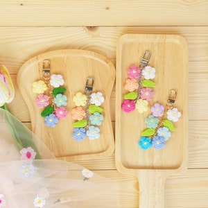 May include: Three crocheted flower keychain charms on wooden trays. The charms are made of colorful yarn and feature a variety of flower shapes. The charms are attached to silver key rings.