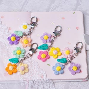 May include: Crocheted flower keychains in various colors, including white, purple, pink, yellow, orange, and blue. Each keychain features multiple flowers, a green leaf, and a silver-tone clasp. The keychains are displayed on a pink notebook.