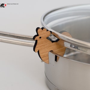 Peut inclure: Un support de couvercle de casserole en bois en forme de coq avec un contour noir. Le support est fixé à la poignée d'une casserole en acier inoxydable avec un couvercle en verre.