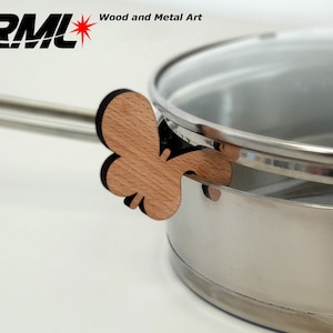 May include: A stainless steel pot with a glass lid and a wooden butterfly-shaped handle. The handle is attached to the pot with a metal hinge. The pot is sitting on a white surface. The text "Wood and Metal Art" is visible on the lid.