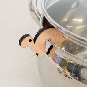May include: A wooden pot lid holder shaped like a stylized animal, with a brown outline, is holding a stainless steel pot lid.