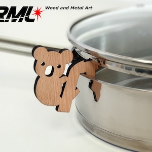 Op de afbeelding: Een houten koalabeer-pannenlap is bevestigd aan een roestvrijstalen pan met een glazen deksel. De pannenlap is lichtbruin van kleur en heeft een zwarte omtrek. De tekst "RML Wood and Metal Art" is zichtbaar in de linkerbovenhoek van de afbeelding.