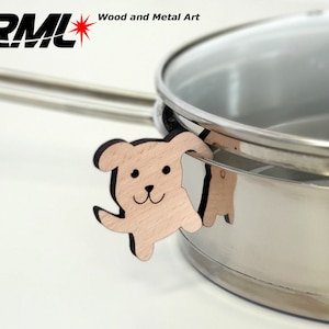 Op de afbeelding: Een roestvrijstalen pan met een glazen deksel en een houten potdeksel in de vorm van een hond. De potdeksel is bevestigd aan de handgreep van de pan. De potdeksel is gemaakt door RML Wood and Metal Art.