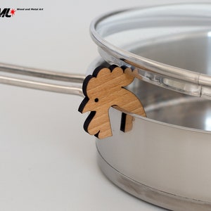 May include: A wooden rooster-shaped pot lid holder in natural wood tones with black accents. The holder is attached to a stainless steel pot with a glass lid.