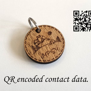 Op de afbeelding: Een houten sleutelhanger met een lasergegraveerd ontwerp van een berglandschap en de tekst "JMPSY". Een QR-code is zichtbaar op de achtergrond, die waarschijnlijk contactgegevens bevat.