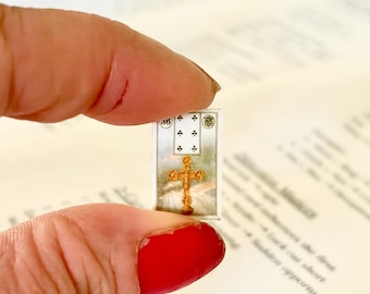 May include: A miniature religious card featuring a gold cross and decorative elements. The card has a rectangular shape and is held between fingers. The background is a blurred view of a printed page.