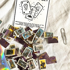 May include: A miniature tarot set with a black and white card design, scattered alongside individual tarot cards. The set includes a paperclip and a card that reads "TINY TAROT SET". The cards have illustrations and a purple and gold design.