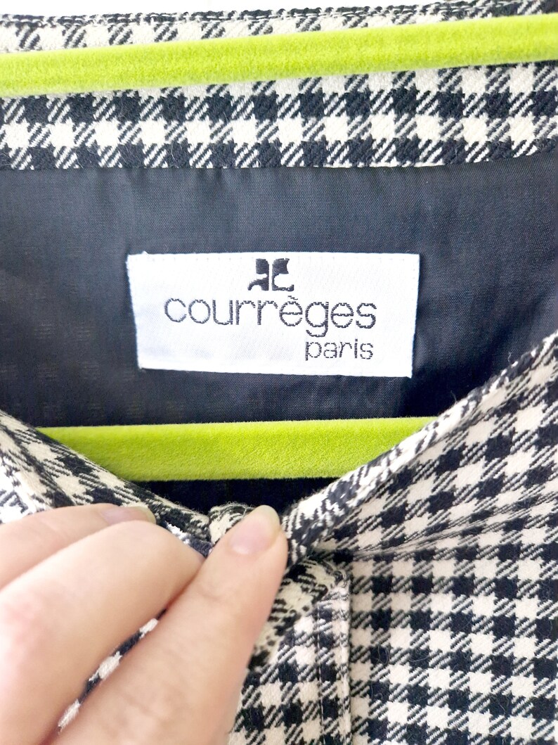 Op de afbeelding: Een shirt van zwart-wit gingham stof met een groene fluwelen hanger en een wit label met de tekst "Courr&egrave;ges Paris".
