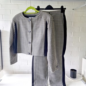 Op de afbeelding: Een zwart-wit geruit broekpak met zwarte bies. Het jasje heeft een ronde hals en knoopsluiting. De broek heeft een rechte pijp.