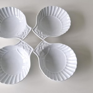 May include: Four white, shell-shaped dishes arranged in a flower-like pattern. Each dish has a ribbed design and a smooth, round centre. The dishes are made of ceramic or porcelain.