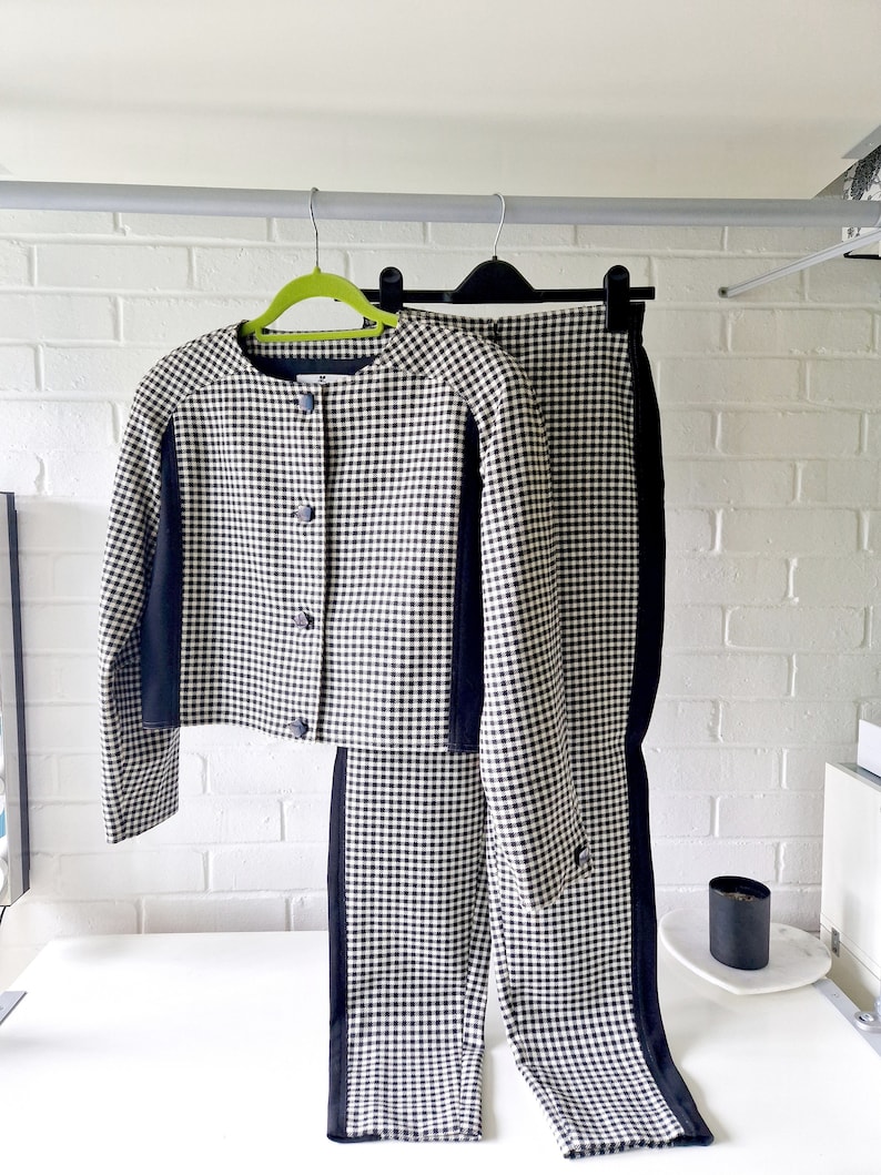 Op de afbeelding: Een zwart-wit geblokt jack en broek set. Het jack heeft zwarte bies aan de mouwen en de broek heeft zwarte bies aan de zijkanten. De set hangt aan een kledingrek.