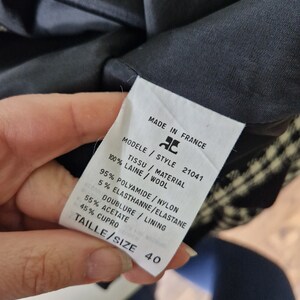 Op de afbeelding: Een wit label met zwarte tekst met details over de stofsamenstelling en de maat van een zwart-wit geruite blazer. Het label vermeldt "Made in France", "Modele Style 21041", "Tissu / Material 100% Laine / Wool", "95% Polyamide / Nylon 5% Elasthanne / Elastane", "Doublure / Lining 55% Acetate 45% Cupro", en "Taille / Size 40".