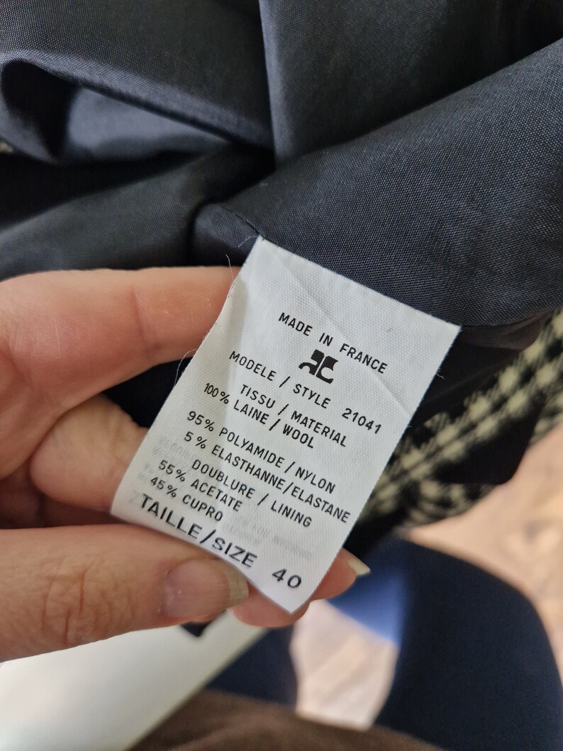Op de afbeelding: Een wit label met zwarte tekst met details over de stofsamenstelling en de maat van een zwart-wit geruite blazer. Het label vermeldt "Made in France", "Modele Style 21041", "Tissu / Material 100% Laine / Wool", "95% Polyamide / Nylon 5% Elasthanne / Elastane", "Doublure / Lining 55% Acetate 45% Cupro", en "Taille / Size 40".
