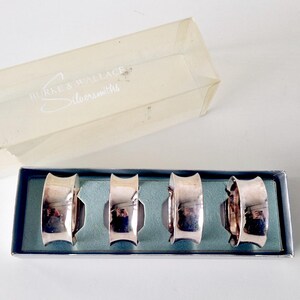 May include: A set of four silver napkin rings presented in a blue box with a clear lid. The napkin rings have a curved design. The box is labelled "Silverworks".