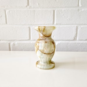 May include: A small, decorative onyx vase with a flared top and base. The vase features a light green and brown marbled pattern. The vase is approximately 5 inches tall and sits on a white surface against a white brick wall.