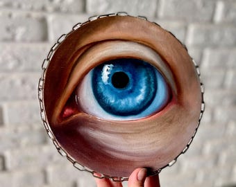May include: A round painting of a realistic blue eye, framed with a silver chain. The eye is detailed with highlights and shadows, set against a neutral skin tone background. The artwork is held by a hand.