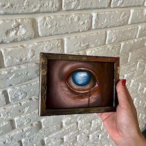 以下が含まれることがあります： 頬を伝う赤い涙を流した、大きく青い目の絵画が額装されています。目は写実的に描かれており、背景は濃い茶色です。