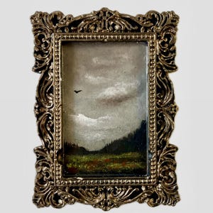 Puede incluir: Una pequeña pintura de paisaje en un marco decorativo dorado. La obra de arte representa un cielo nublado, una línea de árboles oscura y un campo de color verde y rojo. Un pequeño pájaro es visible en el cielo. El marco tiene un diseño intrincado y vintage.