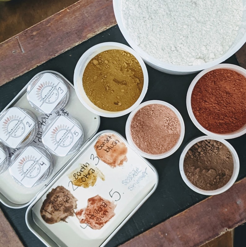 Puede incluir: Un conjunto de seis peque&ntilde;os recipientes de polvos de diferentes colores, etiquetados con n&uacute;meros y nombres como "Earth Okra" y "Sunset Blush". Los recipientes est&aacute;n dispuestos en c&iacute;rculo alrededor de un recipiente m&aacute;s grande de polvo blanco.