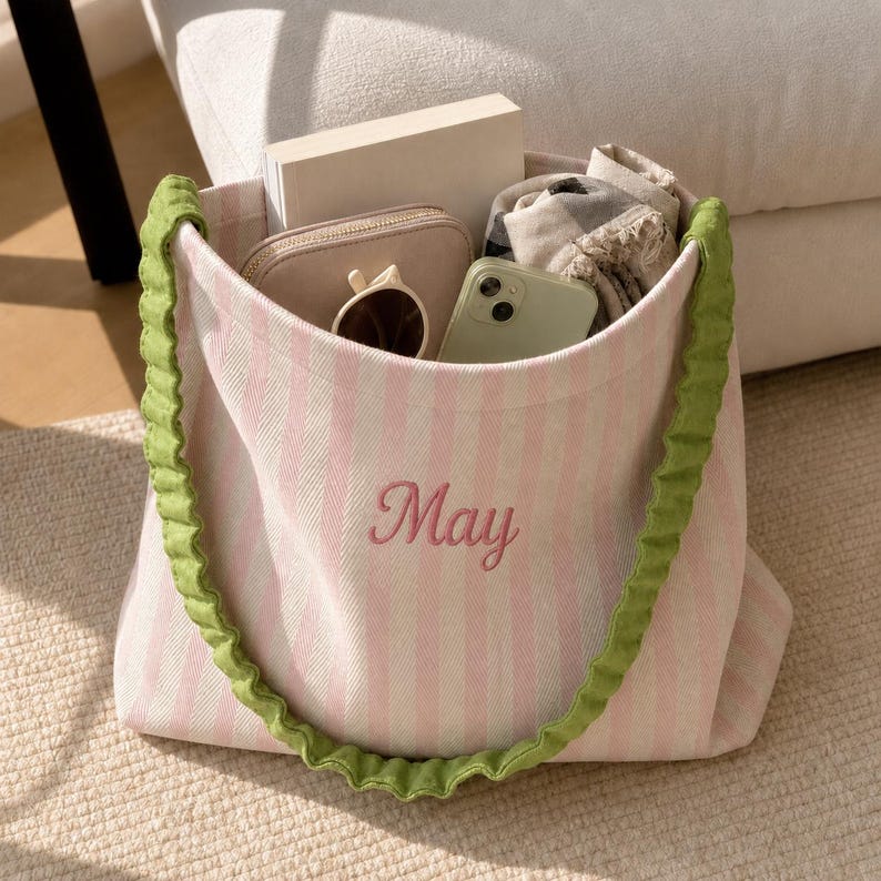 May include: A pink and white striped canvas tote bag featuring the embroidered name May in pink script. The bag has ruffled green fabric straps and contains a white book, a beige zippered pouch, sunglasses, and a smartphone.