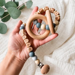 May include: A baby teether set featuring a wooden ring and a pacifier clip. The ring has light blue, cream, and wooden beads with the name DUNCAN engraved. The clip has a wooden end, blue and cream beads, and wooden blocks spelling DUNCAN.