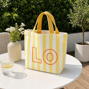 May include: A canvas tote bag featuring vertical yellow and white stripes with two yellow fabric handles. The front of the bag displays the letters LO in a bold orange outline. The bag has a rectangular shape and is constructed from fabric.