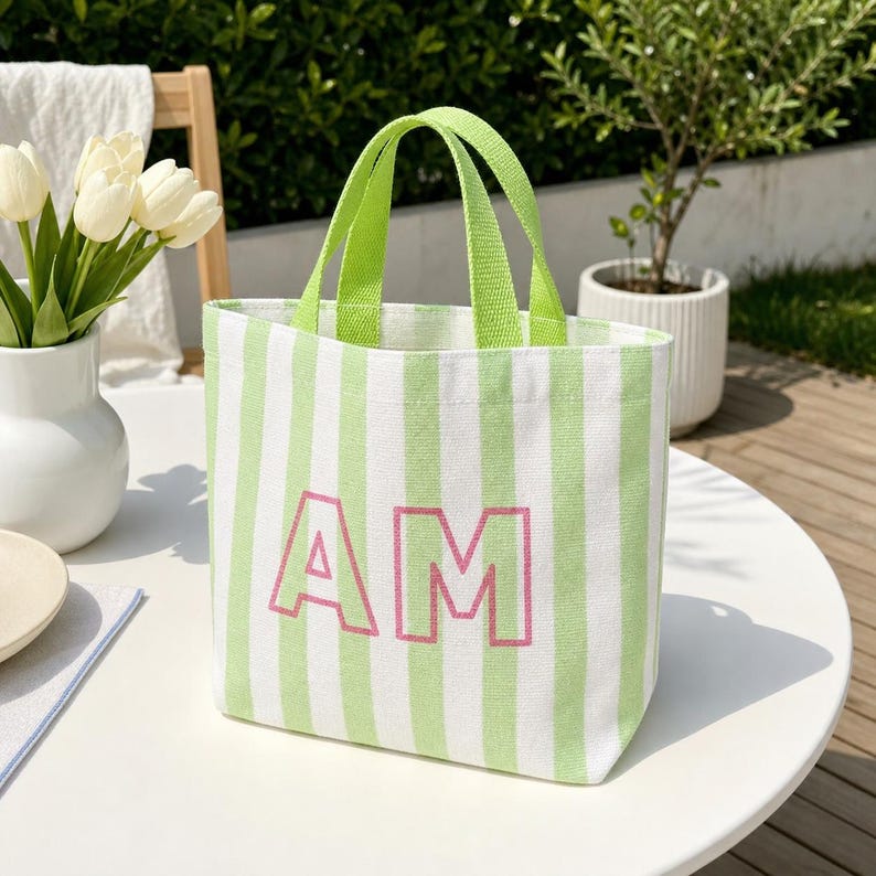 May include: A canvas tote bag featuring vertical light green and white stripes with two lime green fabric handles. The front of the bag displays the letters AM in a bold, pink, outlined font centered on the striped pattern.