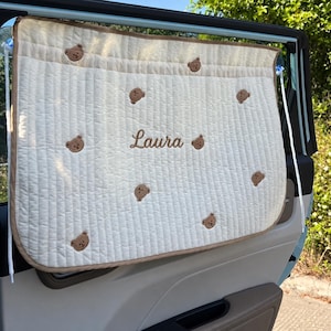 May include: A cream-colored quilted car window shade featuring a repeating pattern of small brown embroidered teddy bear faces and the name Laura stitched in brown thread in the center, secured with white cords and clear suction cups.